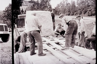 Group of men unload a large stone slab from a truck bed. Workers in work clothes and hats appear to be in a construction sett...