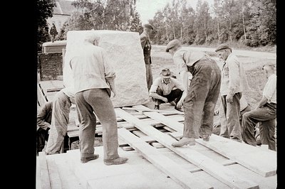 Men in workwear and hats load large, pale stone blocks onto a flatbed truck. Possible construction or quarrying scene. Weathe...