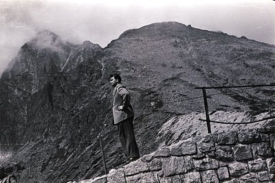 Man in a suit stands atop a stone wall overlooking a dramatic, mist-shrouded mountain range. Likely a travel or resort scene,...