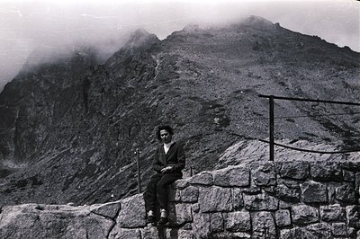 A seated individual rests on a stone wall overlooking a steep, mist-shrouded mountain landscape. The subject wears trousers a...