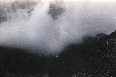 Dramatic, monochrome landscape depicting rugged, dark cliffs rising to meet stormy skies. Visible image degradation—likely sc...