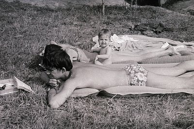 Several people sunbathe on grass, with a baby positioned centrally. A woman in a floral swimsuit lies on a blanket near the e...
