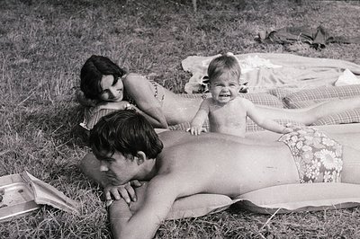 A family portrait – a man sunbathing, a woman reclined, and a toddler playfully posed on a patterned blanket. Likely a candid...