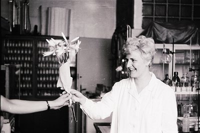 A smiling woman in a lab coat receives a bouquet wrapped in paper. The laboratory setting shows rows of bottles and scientifi...