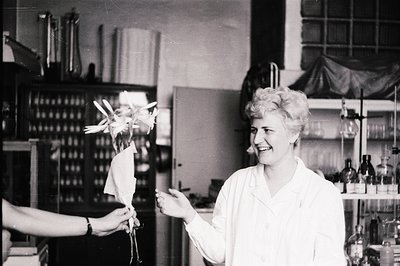 A woman in a lab coat smiles while presenting a pinwheel. Shelves with bottles & glassware dominate the background, suggestin...