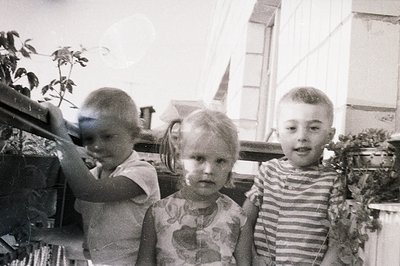 Three children stand behind a railing on a balcony, likely in a seaside setting. The image, grainy and exhibiting surface wea...