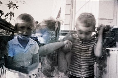 A poignant snapshot of three children outdoors. Two boys, one holding a girl, appear in mid-motion, likely playing. The grain...