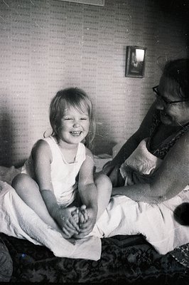 A young child, seated on a bed covered with a patterned bedspread, smiles widely. A woman sits beside the child, partially ob...