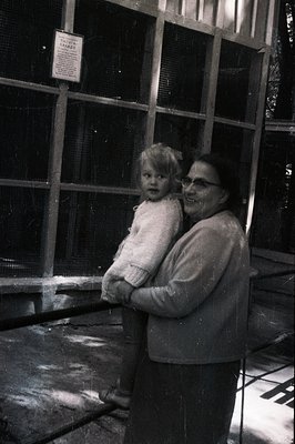 A smiling woman carries a young child in her arms, both bundled in sweaters. The scene features a wire mesh enclosure, possib...