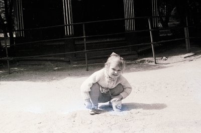 A young child, likely a toddler, kneels on a sandy surface, appearing to examine the ground. They wear a textured sweater and...