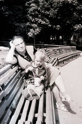 A seated adult and a young child rest on a long, slatted park bench. The adult, wearing a short-sleeved shirt and shorts, has...