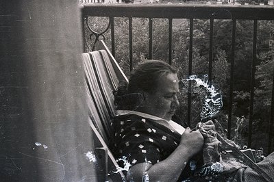 A woman sits in a wrought-iron chair, engaged in knitting. She wears a dark, dotted dress and a scarf. The background feature...