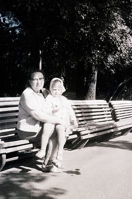 A woman holds a young child on a long park bench beneath mature trees. Both wear vintage attire, suggesting a 1960s or 1970s ...