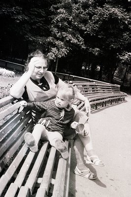 A young boy sits on a woman’s lap on a park bench. She holds him close, gently touching his face; both appear to be enjoying ...