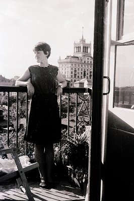 A woman stands on a balcony overlooking a grand, ornate building, likely a seaside resort. She wears a dark, sleeveless dress...