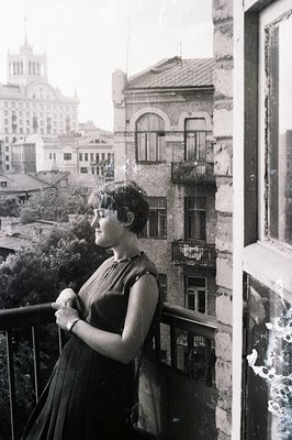 Young woman in a dark dress poses on a balcony overlooking a cityscape. Notable architecture suggests Eastern European influe...