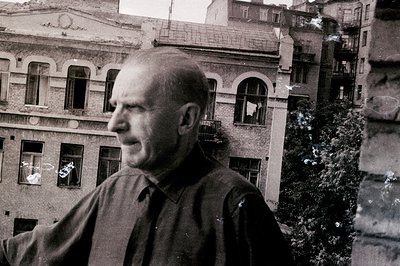 A middle-aged man with a bald head is seen in profile, appearing contemplative against a backdrop of aging European architect...