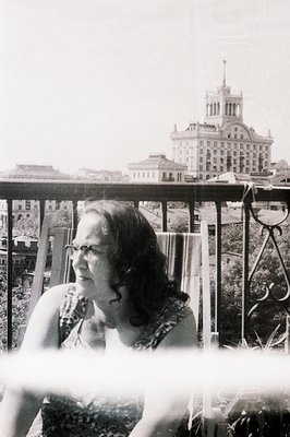A woman with shoulder-length curly hair gazes off-camera from behind ornate ironwork, with a grand, multi-tiered building vis...