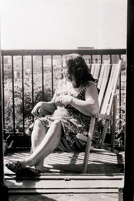 A woman in a floral dress sits in a wooden rocking chair on a balcony overlooking a cityscape. She wears glasses and appears ...
