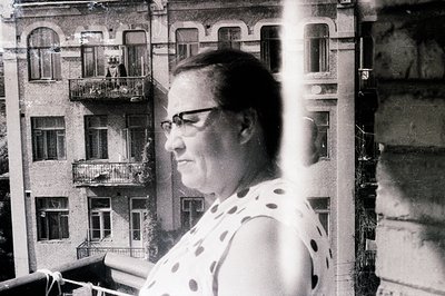 A woman in spectacles stands on a balcony, gazing outwards. A brick building with ornate balconies forms the backdrop. The im...