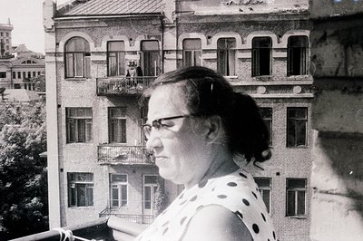A side-profile portrait of a woman in a polka-dot dress and eyeglasses, framed by a classical-style building facade. The arch...