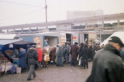 A snowy, bustling scene featuring a public market beneath elevated train tracks. People bundled in winter coats crowd around ...