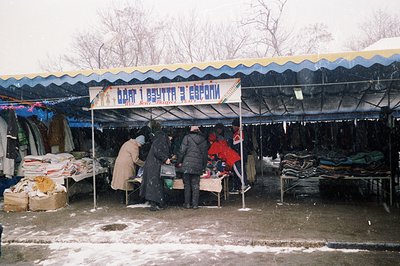 A snowy outdoor market scene. Several people, bundled in winter clothing, browse tables piled high with shoes, coats, and oth...