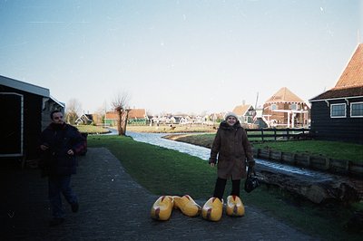 A canal-side scene in a Dutch village. A woman in a brown coat stands smiling near oversized wooden clogs; a man runs towards...