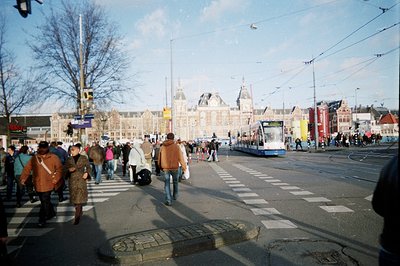 A bustling scene at a city intersection, likely in the Netherlands. A tram passes pedestrians crossing the street in front of...
