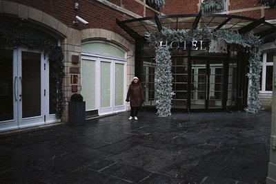 A person stands outside a hotel entrance, sheltered by a modern awning decorated with silver foliage. Wet paving stones sugge...