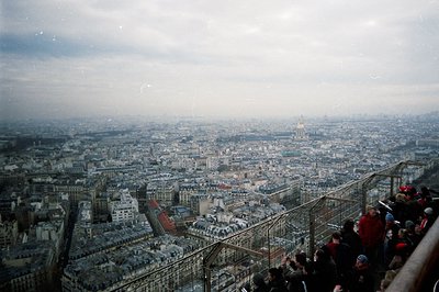 Panoramic view of Paris rooftops under a hazy sky, featuring the dome of Sacré-Cœur Basilica prominently visible. A crowd of ...