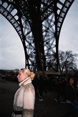 A woman in a fur coat stands in profile against the backdrop of the Eiffel Tower. The grainy, cool-toned image suggests a 199...