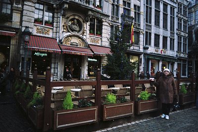 A bustling scene on cobblestone street featuring a cafe with outdoor seating & a decorated Christmas tree. Ornate, historic a...