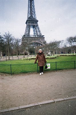 A woman stands facing the Eiffel Tower, framed by a low wire fence and park landscaping. She wears a fur-trimmed coat, dark p...