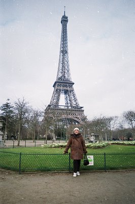 A woman stands in front of the Eiffel Tower, framed by a wire fence and a grassy park. The tower dominates the background und...