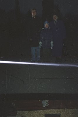 Three figures stand on a rooftop terrace overlooking a city at night. The adults are dressed in dark coats, while a younger i...