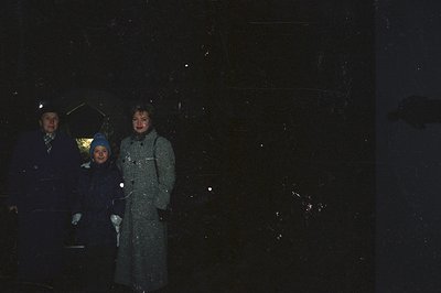 A man, woman, and child stand outdoors in what appears to be a rain shower. The man holds an umbrella; the child wears a brig...