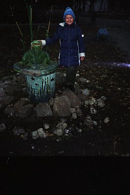 A young child, bundled in a blue coat and hat, stands next to a large, ornate frog statue. The statue sits on a tiered base s...