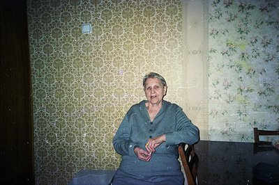 An older woman sits in a dining chair, facing the camera. She wears a gray cardigan and has short, styled gray hair. Backgrou...