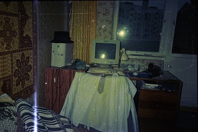 A vintage desktop computer setup occupies a small, cluttered room. A monitor, tower, and keyboard sit atop a dresser draped w...