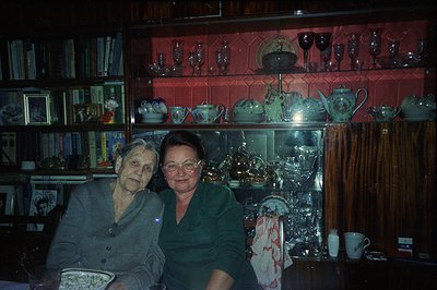 Two women seated at a table in a richly decorated dining room, bookshelves flanking a large display cabinet filled with china...