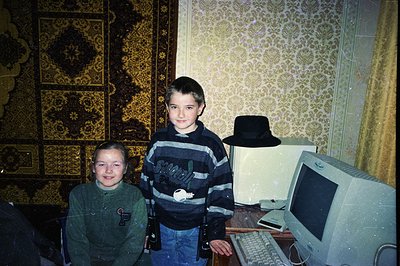 Two boys stand before a vintage beige computer setup in a room with patterned wallpaper and a patterned rug. Both wear casual...