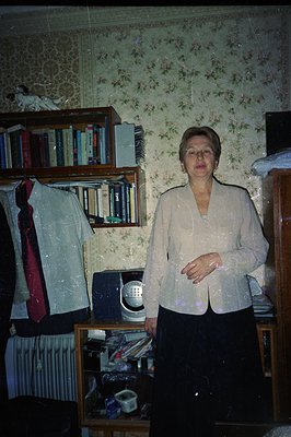A mature woman stands in a room with wallpaper patterned with floral motifs. She wears a light-colored suit jacket over a dar...