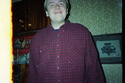 A man in a red and black plaid shirt stands in what appears to be a vintage dining room. Dark wood cabinetry and a large, orn...