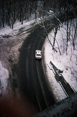 Overhead shot: a dark-colored car navigates a winding, snow-covered road cutting through a dense forest. Visible road marking...