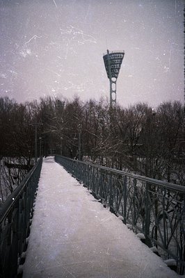 A snowy, pedestrian bridge leads towards a tall stadium floodlight tower amidst bare trees. A frozen river or body of water i...