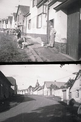 A village street scene; traditional architecture with steep-roofed houses visible. Foreground depicts a man in workwear and a...