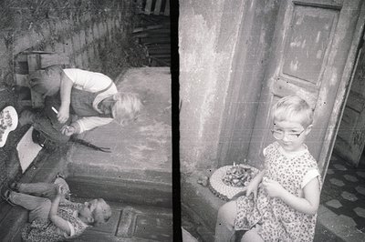 Split-tone, b&w photo. A girl in a dress & glasses sits beside a basket of berries; another girl appears to be playing with a...