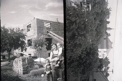 A partially constructed brick house under development. A woman sits with two young children, seemingly observing the construc...