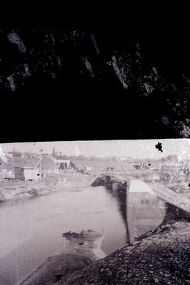 Monochrome image depicting a bridge over a waterway. Visible are industrial structures, likely a harbor or port, with a dense...